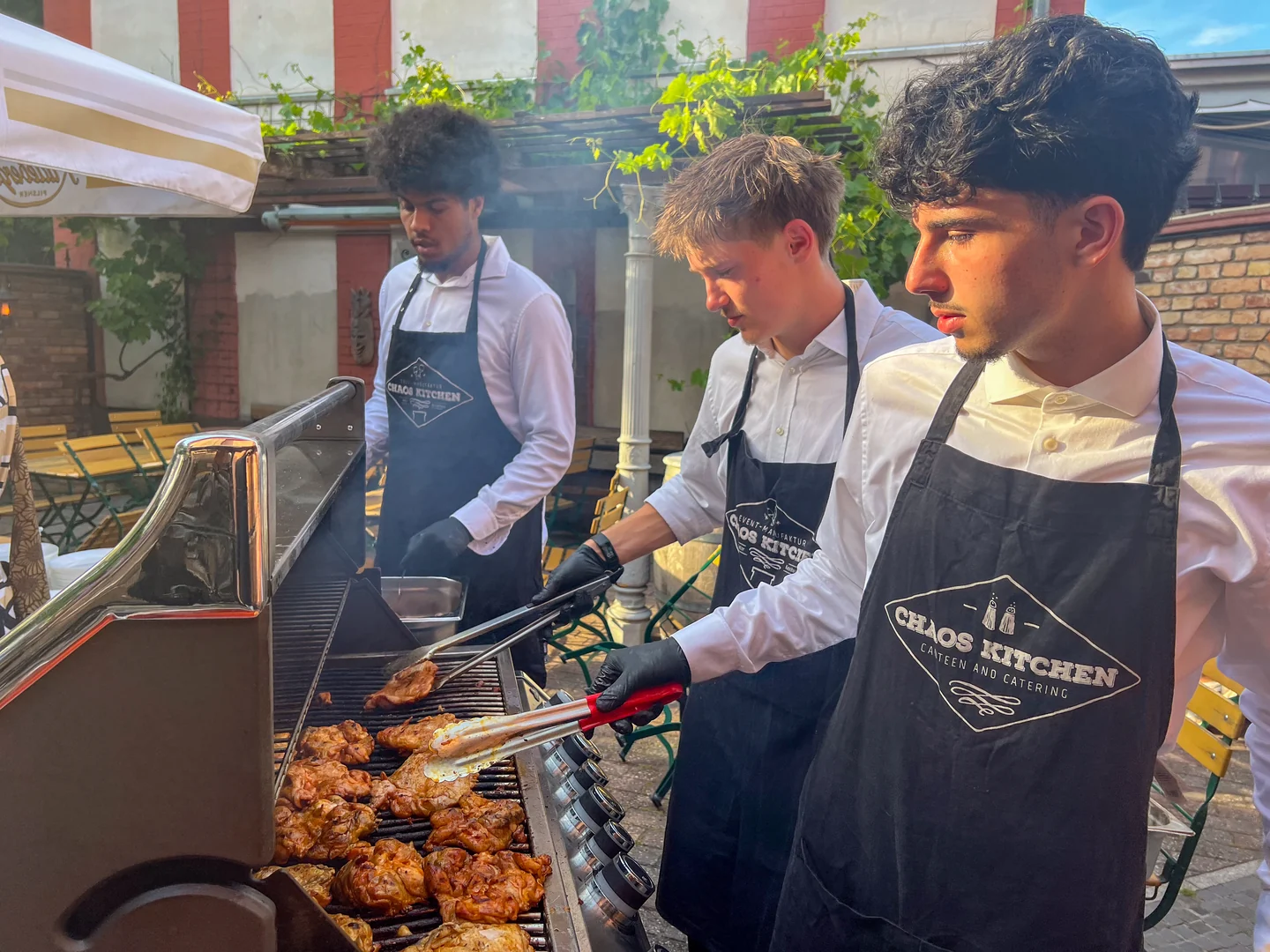 Drei Personen mit Chaos-Kitchen-Schürzen arbeiten am Grill im Hof des XELOR Kesselhauses.