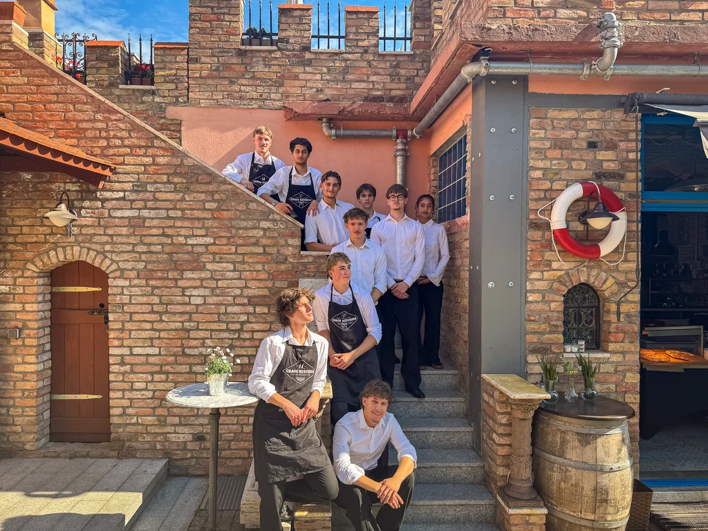 Service-Team von Chaos Kitchen in weißen Hemden und schwarzen Schürzen auf der Backsteintreppe des XELOR Kesselhauses in Berlin-Neukölln.